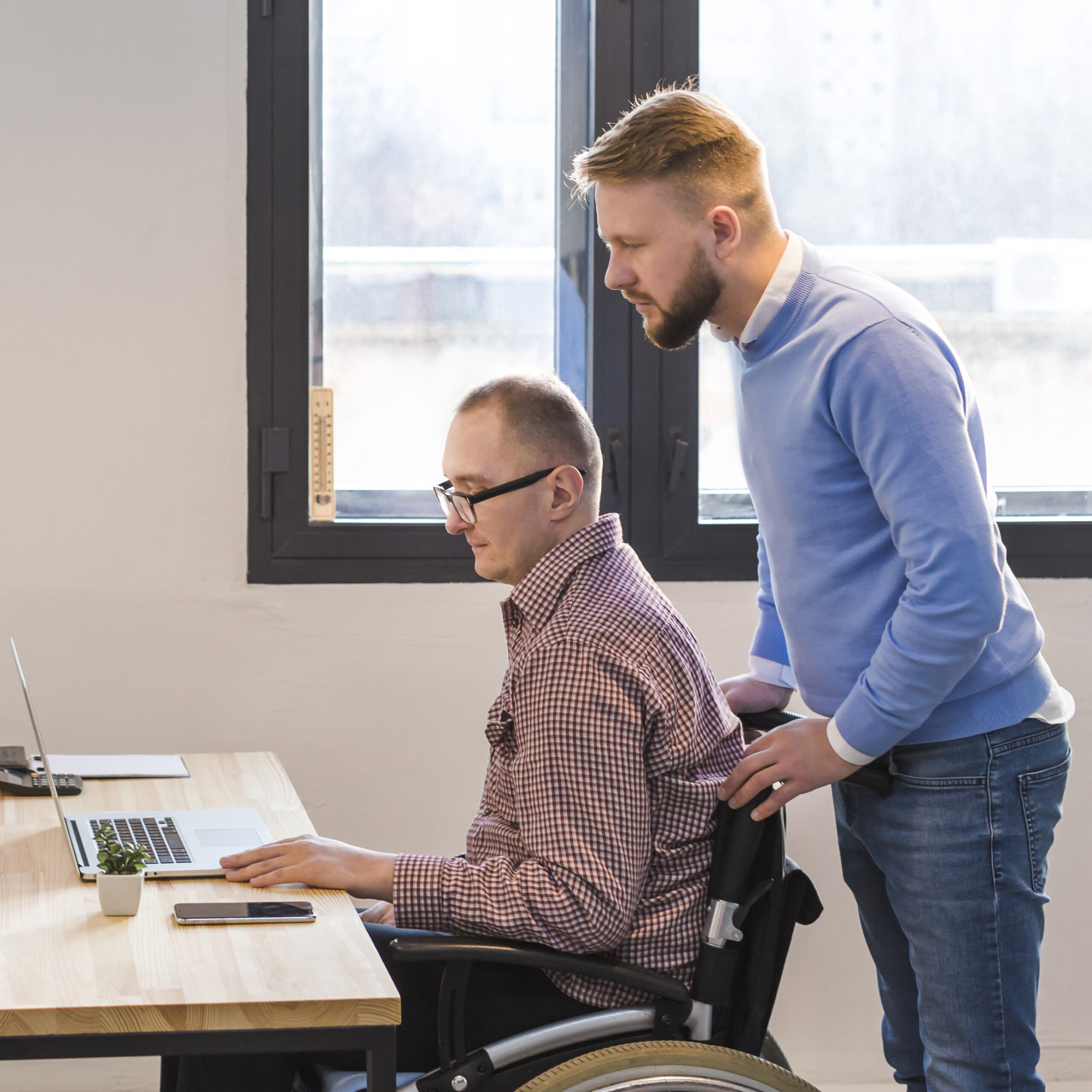 A wheelchair person at a desk working with another person stood behind them leaning onto the back of the wheelchair.