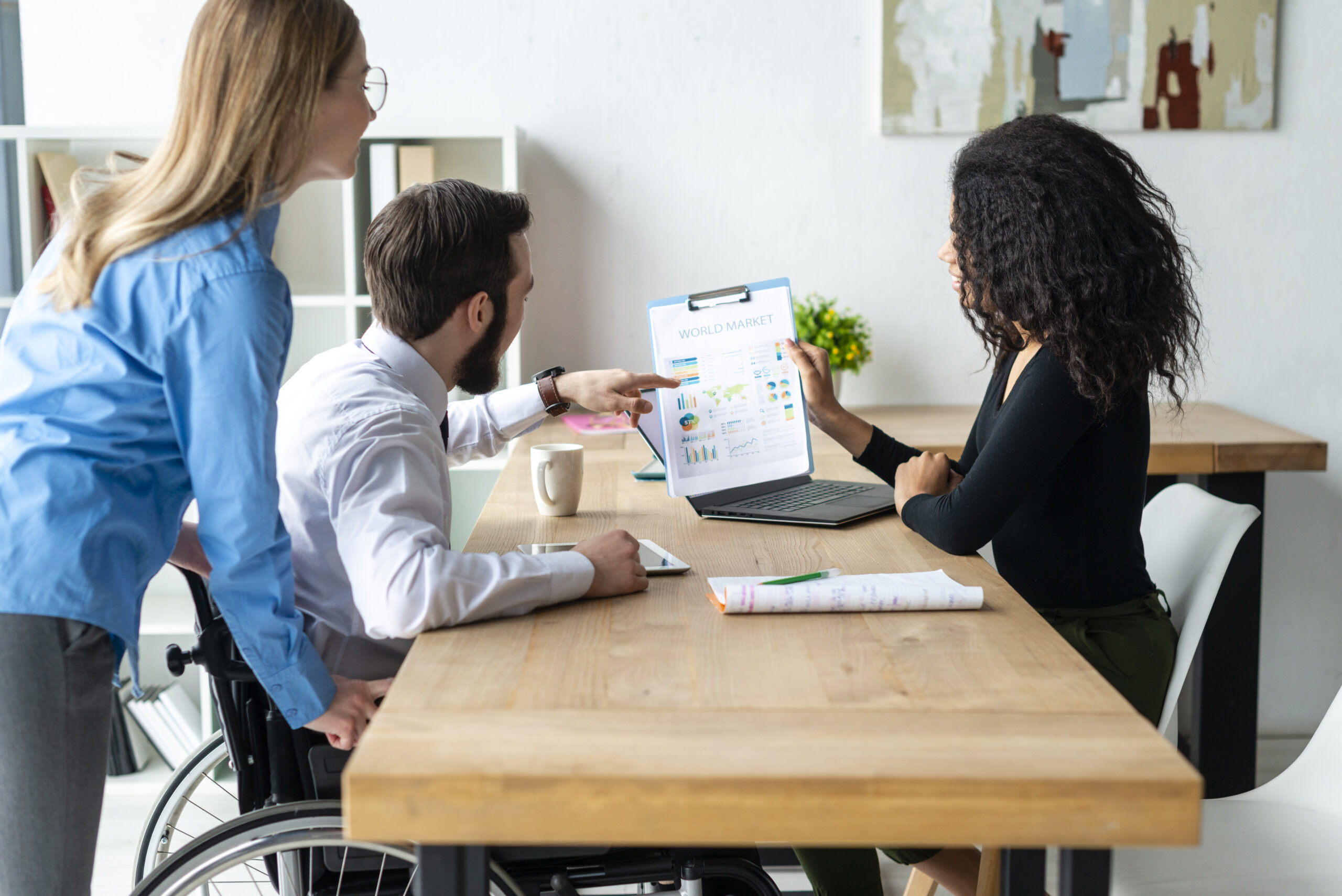 Three people working in an office around a desk, one person sat on a chair showing two others work on a laptop. One of the people are in a wheelchair and the other person is leaning over the desk.