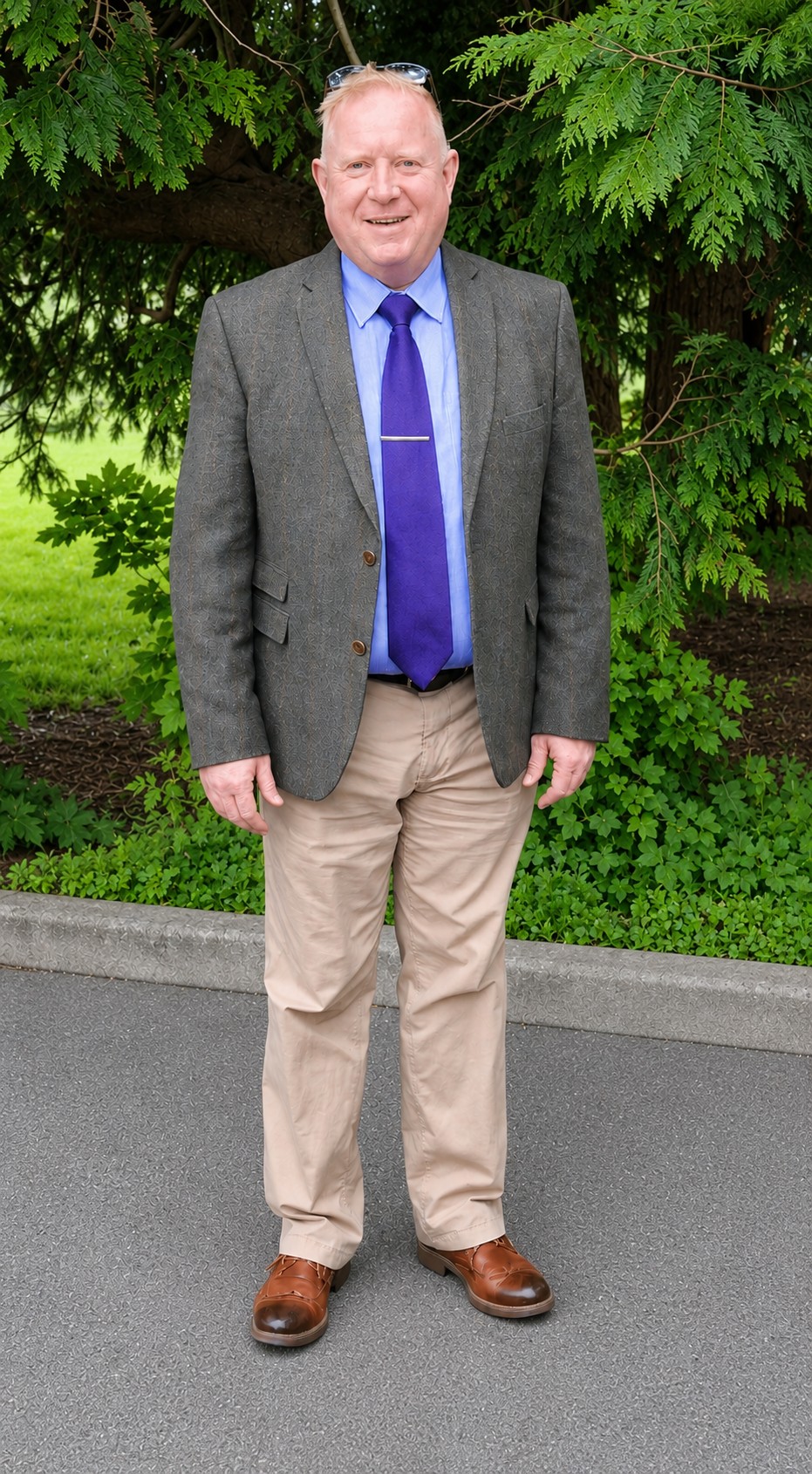Jamie Hanlon, stood in a car park with a green tree in the background, grass and foliage. Jamie is wearing a tweed jacket, light blue shirt, purple tie, beige trousers and brown shoes. 